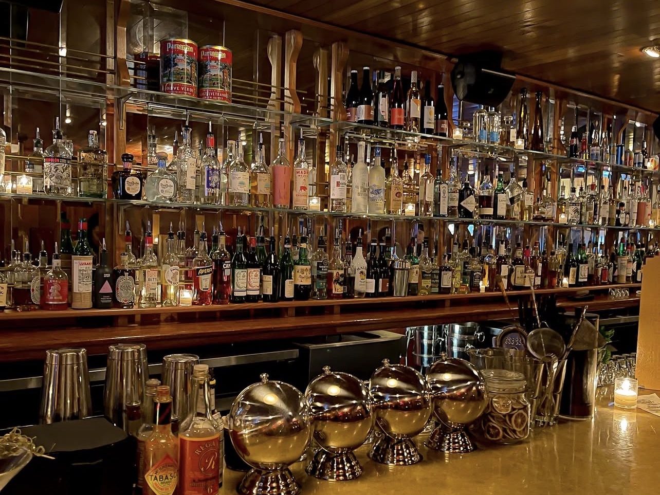 A well-stocked bar with an extensive selection of spirits on shelves, featuring various bottles and mixers, illuminated by a warm overhead light.